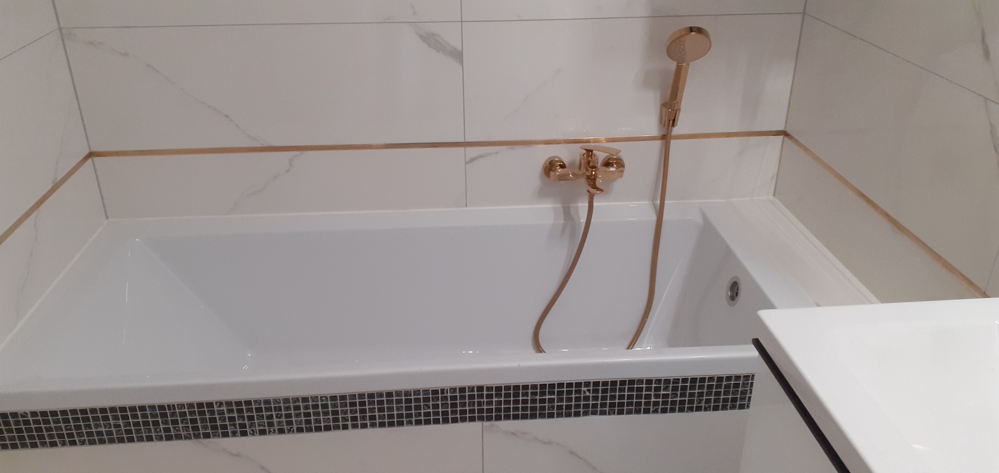 Biała wanna obudowana mozaiką, złota armatura i białe marmurowe płytki z delikatnym złotym akcentem tworzą eleganckie wykończenie łazienki.