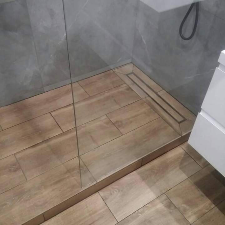 Wykończona kabina prysznicowa walk-in z drewnopodobnymi płytkami na podłodze i szarymi płytkami imitującymi marmur na ścianach, z odpływem liniowym w rogu.