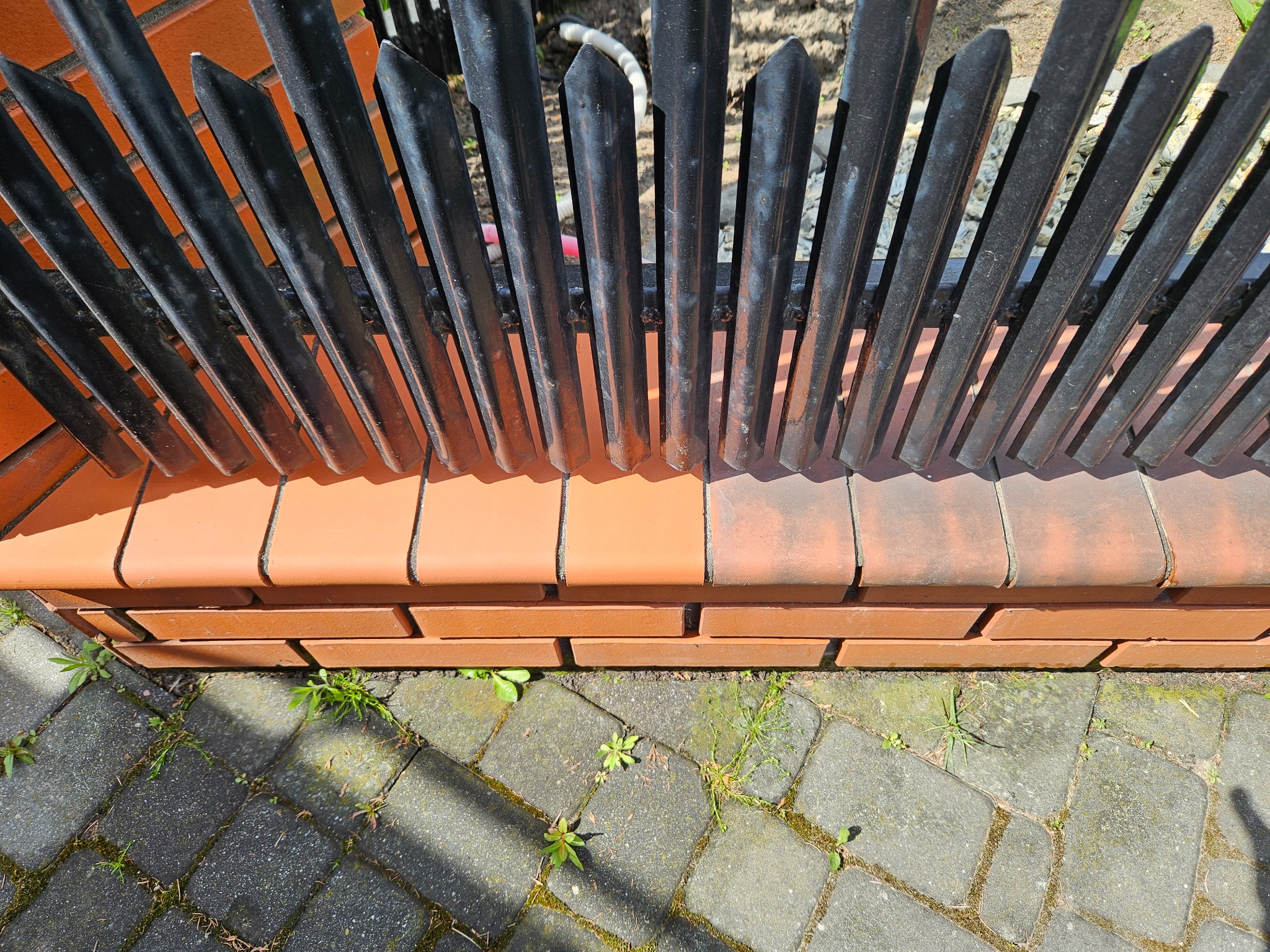 Metalowe ogrodzenie z ostrymi zakończeniami, osadzone na ceglanym murku, z kostką brukową i chwastami u podstawy. Widok z góry, ostre światło słoneczne.