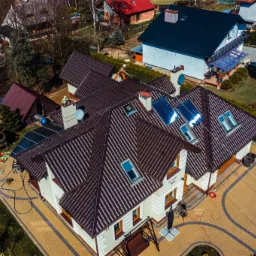 Widok z drona na duży dom z brązowym dachem, pracownicy na dachu zabezpieczeni linami, niebieski basen w ogrodzie, kostka brukowa na podjeździe.