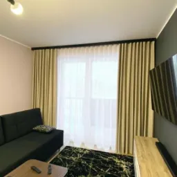 Beżowe zasłony zaciemniające z czarnym karniszem w nowoczesnym salonie z czarną sofą i dywanem w złote żyły, widok z boku na stolik kawowy z pilotami.