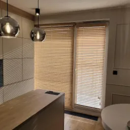 Jasne drewniane żaluzje poziome w nowoczesnej kuchni z szarymi szafkami i okrągłymi lampami wiszącymi z odbijającym światło kloszem.