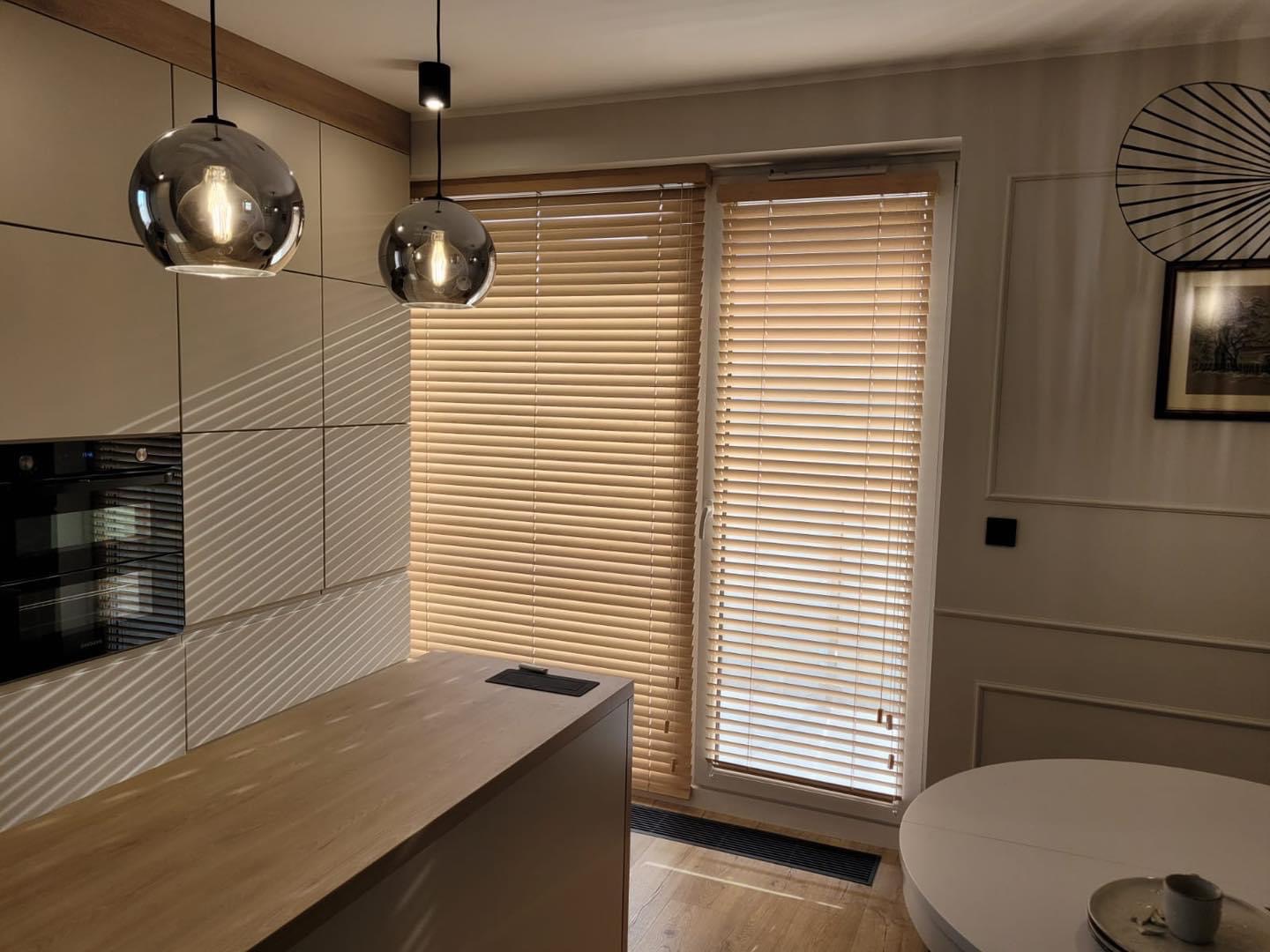 Jasne drewniane żaluzje poziome w nowoczesnej kuchni z szarymi szafkami i okrągłymi lampami wiszącymi z odbijającym światło kloszem.