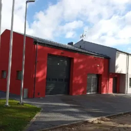 Czerwono-biały budynek straży pożarnej z zamkniętą, segmentową bramą garażową, widok z zewnątrz w słoneczny dzień.