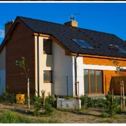 Nowoczesny dom jednorodzinny z czarnym dachem, drewnianą elewacją i oknami połaciowymi, otoczony młodymi drzewkami i ogrodzeniem z siatki.