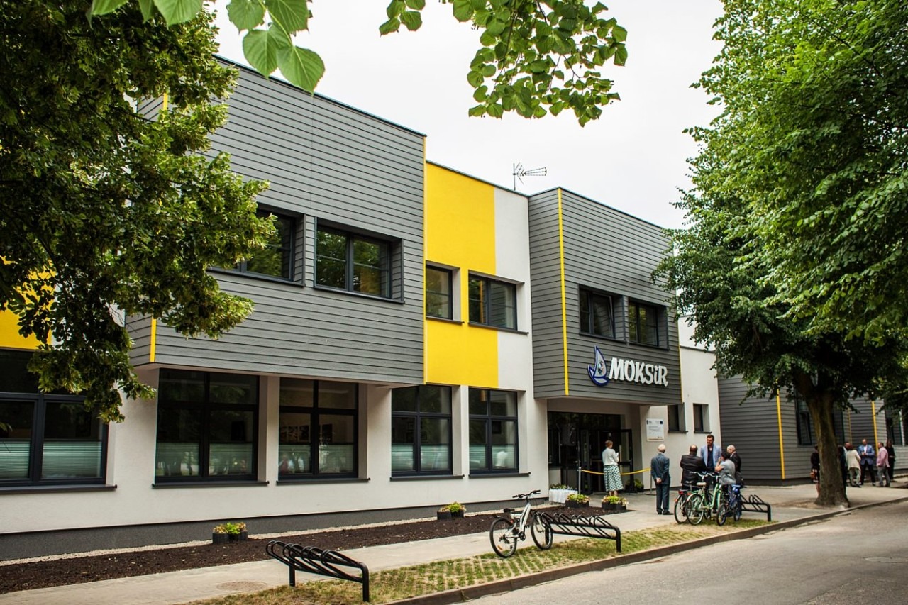Nowoczesny budynek z szarymi panelami elewacyjnymi, żółtymi akcentami i ciemnymi oknami, przed wejściem stoją rowery, w tle widać grupę ludzi.