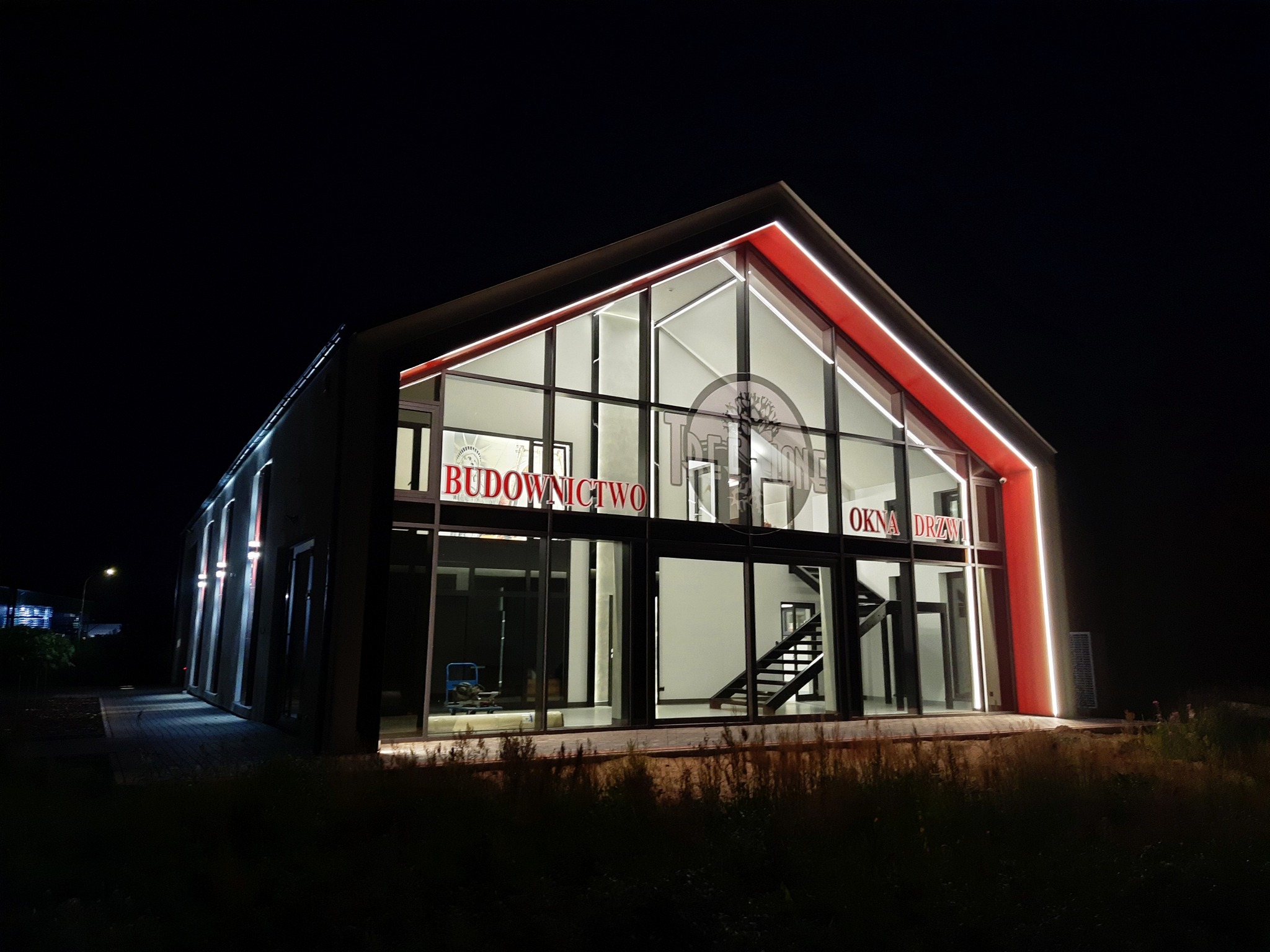 Nowoczesny budynek firmy budowlanej oświetlony nocą, z dużymi przeszkleniami i napisem 'Budownictwo Okna Drzwi' na fasadzie.