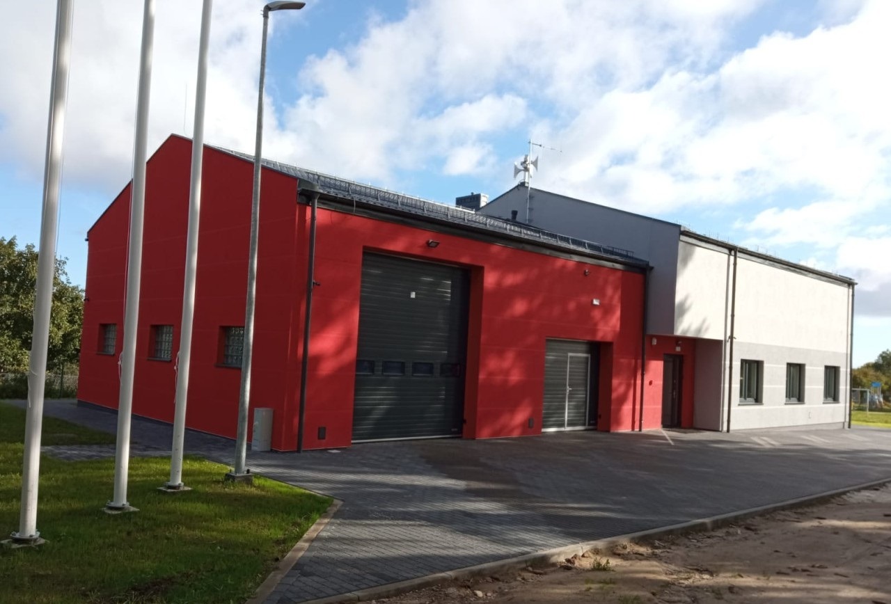 Czerwono-biały budynek straży pożarnej z zamkniętą, segmentową bramą garażową, widok z zewnątrz w słoneczny dzień.