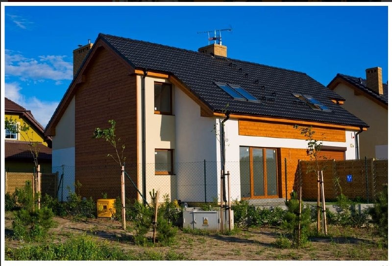 Nowoczesny dom jednorodzinny z czarnym dachem, drewnianą elewacją i oknami połaciowymi, otoczony młodymi drzewkami i ogrodzeniem z siatki.