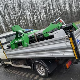 F.U.JARBIR Jarzębowska Bogusława Usługi minikoparką - Ciężarówka z platformą załadowaną zielonym urządzeniem do robót ziemnych marki Simply E70, widok z boku na tle drzew bez liści