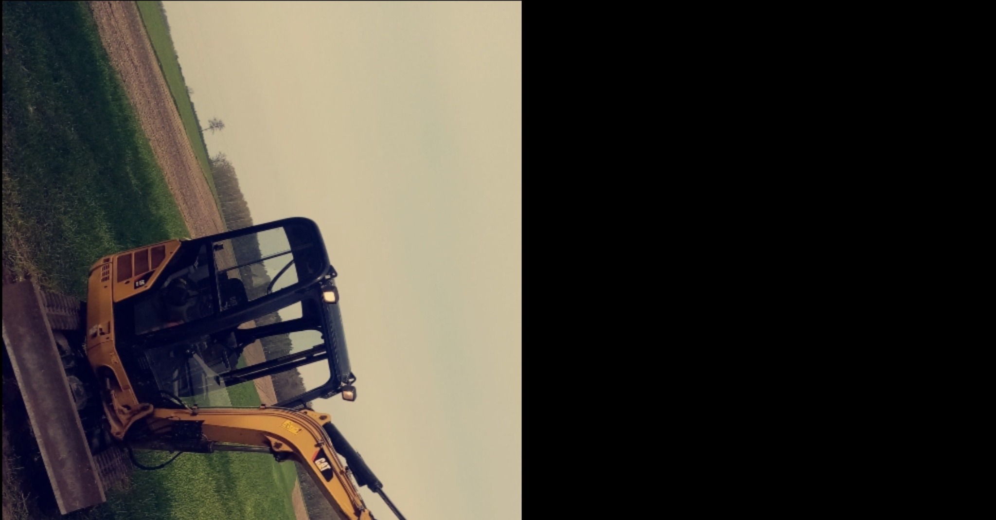 Żółta mini koparka CAT stoi na trawiastym terenie, w tle pole uprawne i pochmurne niebo. Zdjęcie zrobione pod nietypowym kątem.