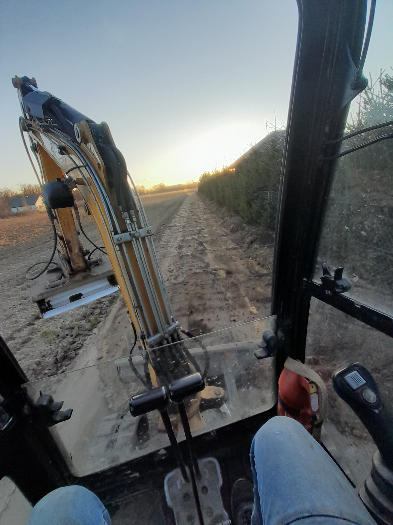 Wnętrze kabiny koparki podczas pracy w polu, widok na ramię koparki i wykopany rów w perspektywie operatora, zachodzące słońce w tle.