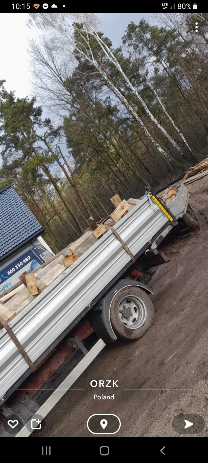 Przyczepa samochodowa z załadunkiem drewnianych elementów, zabezpieczonych pasami transportowymi, na tle lasu i fragmentu dachu budynku.