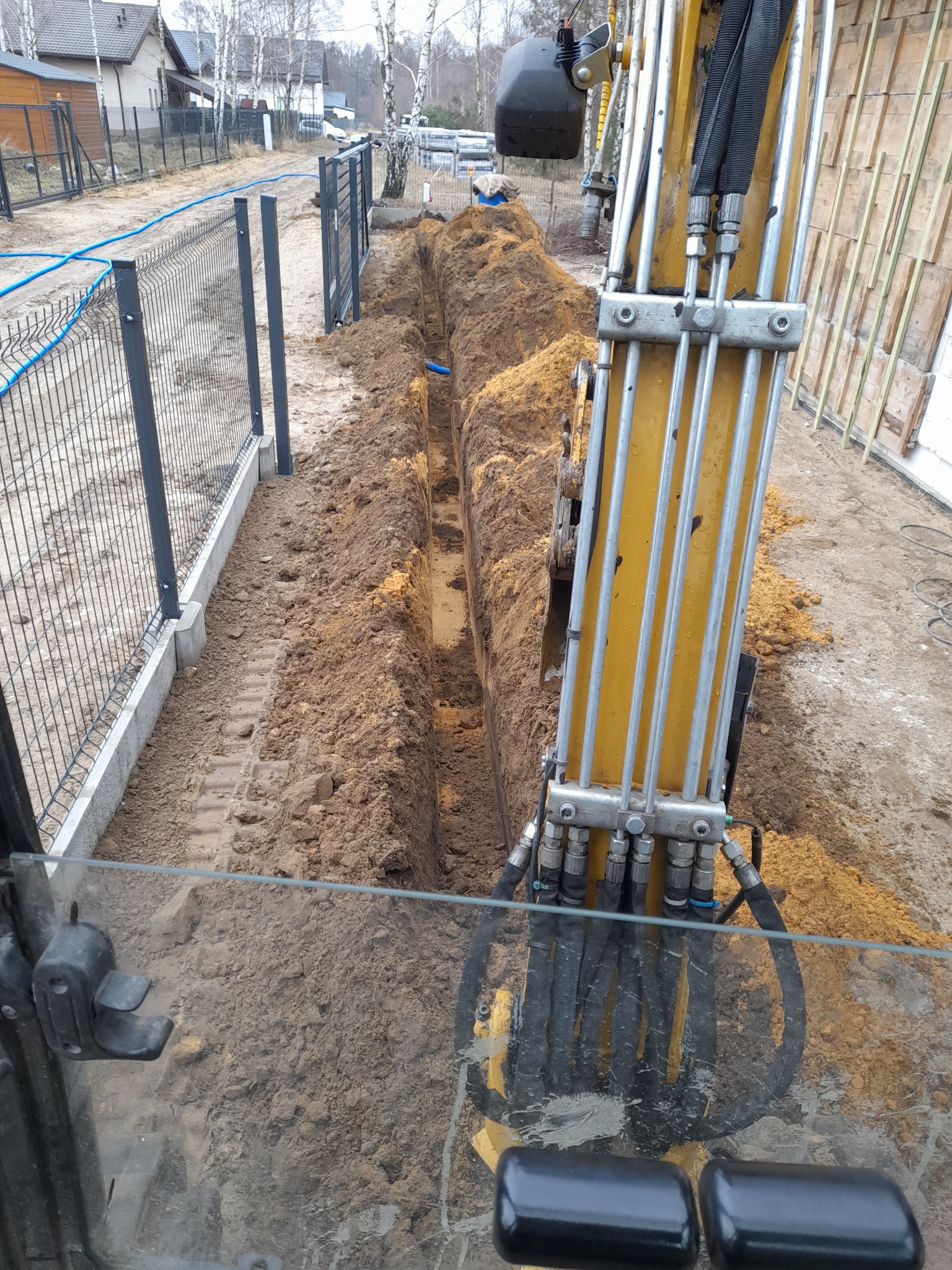 Głęboki, wąski wykop w ziemi, widziany z kabiny żółtej koparki, z widocznymi przewodami hydraulicznymi i fragmentem szyby kabiny; w tle ogrodzenie panelowe i zabudowa jednorodzinna.