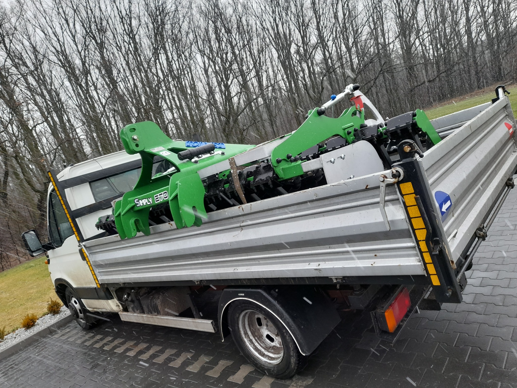 Ciężarówka z platformą załadowaną zielonym urządzeniem do robót ziemnych marki Simply E70, widok z boku na tle drzew bez liści