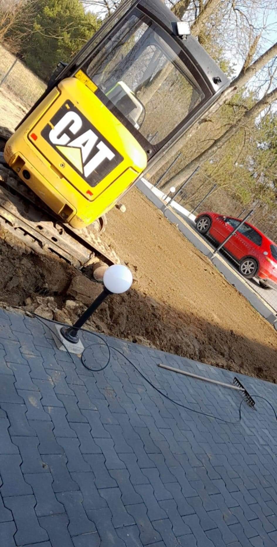 Żółta minikoparka CAT na placu budowy, obok ułożona kostka brukowa, lampa ogrodowa i grabie, w tle czerwony samochód osobowy.
