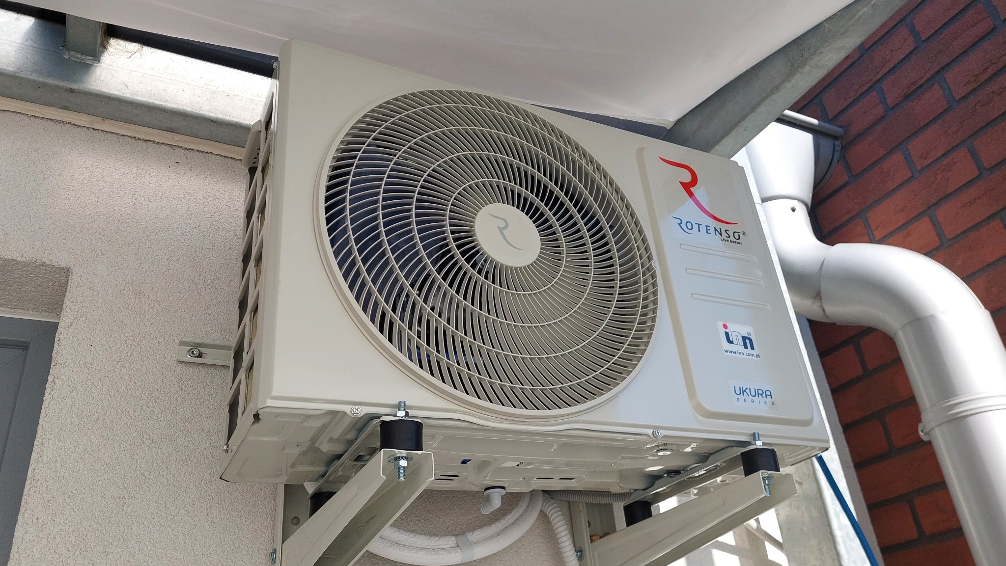 Zewnętrzna jednostka klimatyzacji Rotenso Ukura Series zamontowana na ścianie budynku z cegły, widoczne rury odprowadzające skropliny i elementy mocujące.