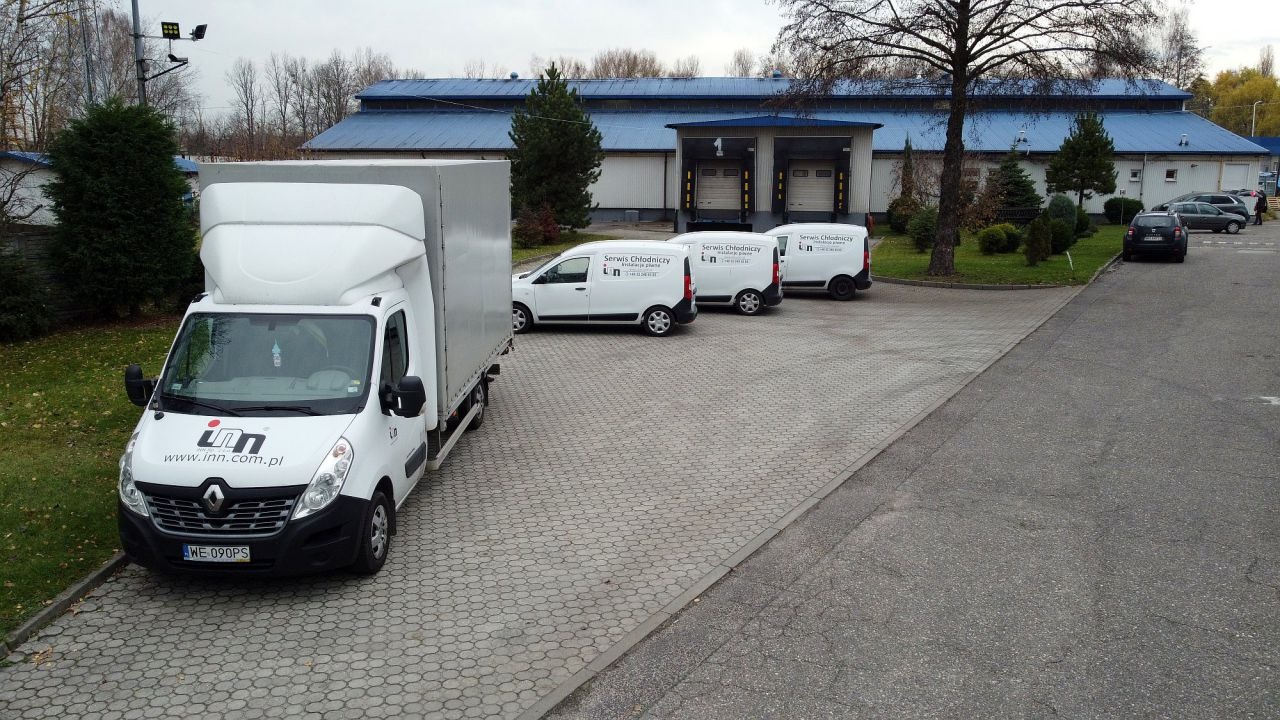 Flota białych samochodów dostawczych z logo firmy serwisującej chłodnictwo, zaparkowana na placu przed budynkiem magazynowym z niebieskim dachem i rampami załadunkowymi.