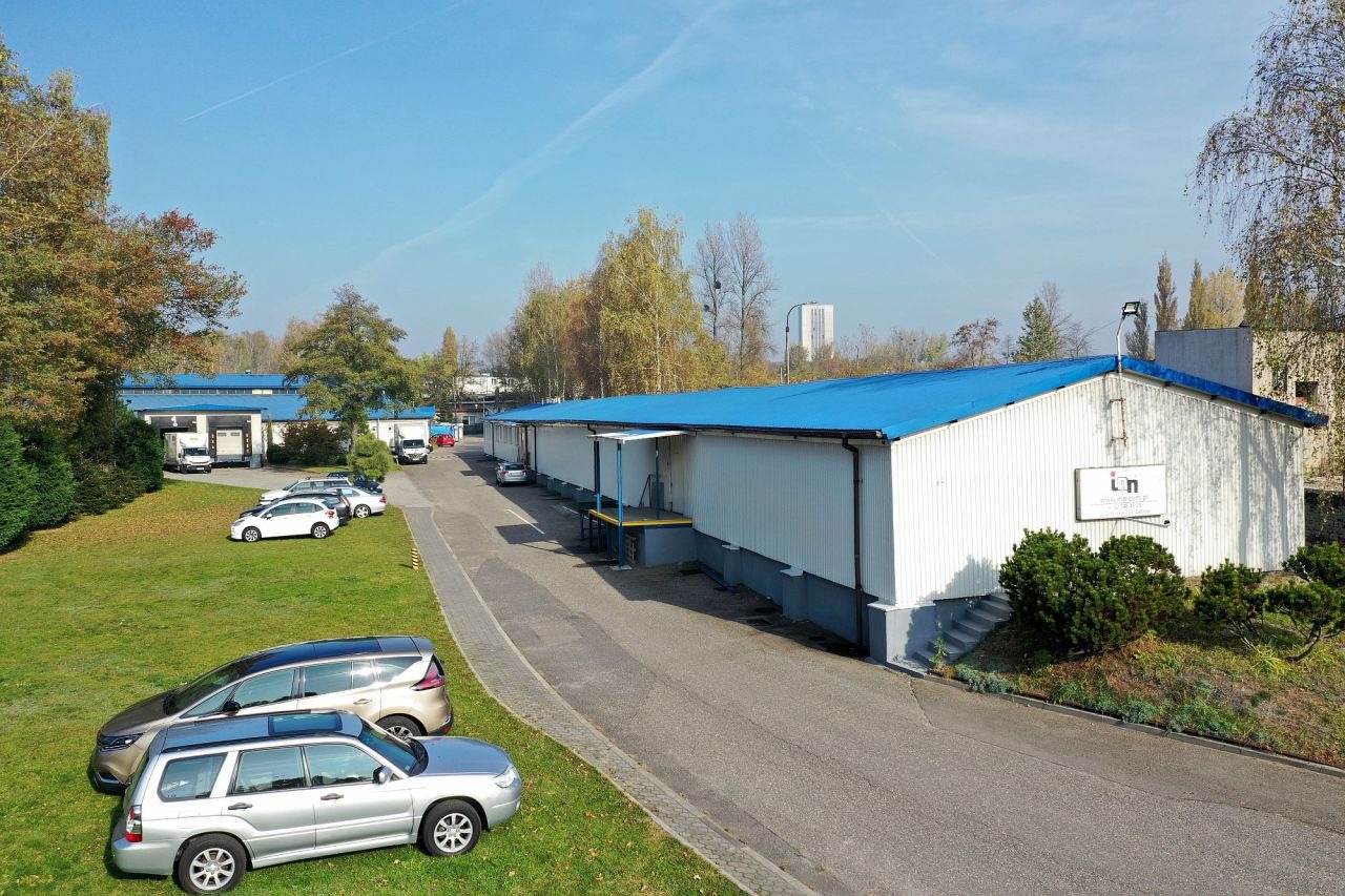 Widok z góry na budynek biurowy z niebieskim dachem, zaparkowanymi samochodami i zielonym trawnikiem w słoneczny dzień.