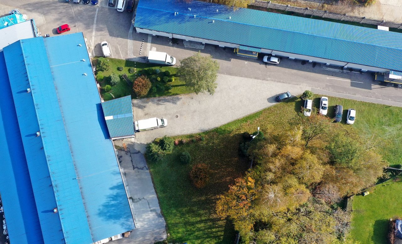 Zdjęcie z lotu ptaka przedstawiające kompleks budynków przemysłowych z niebieskimi dachami, parkingiem i zielenią.