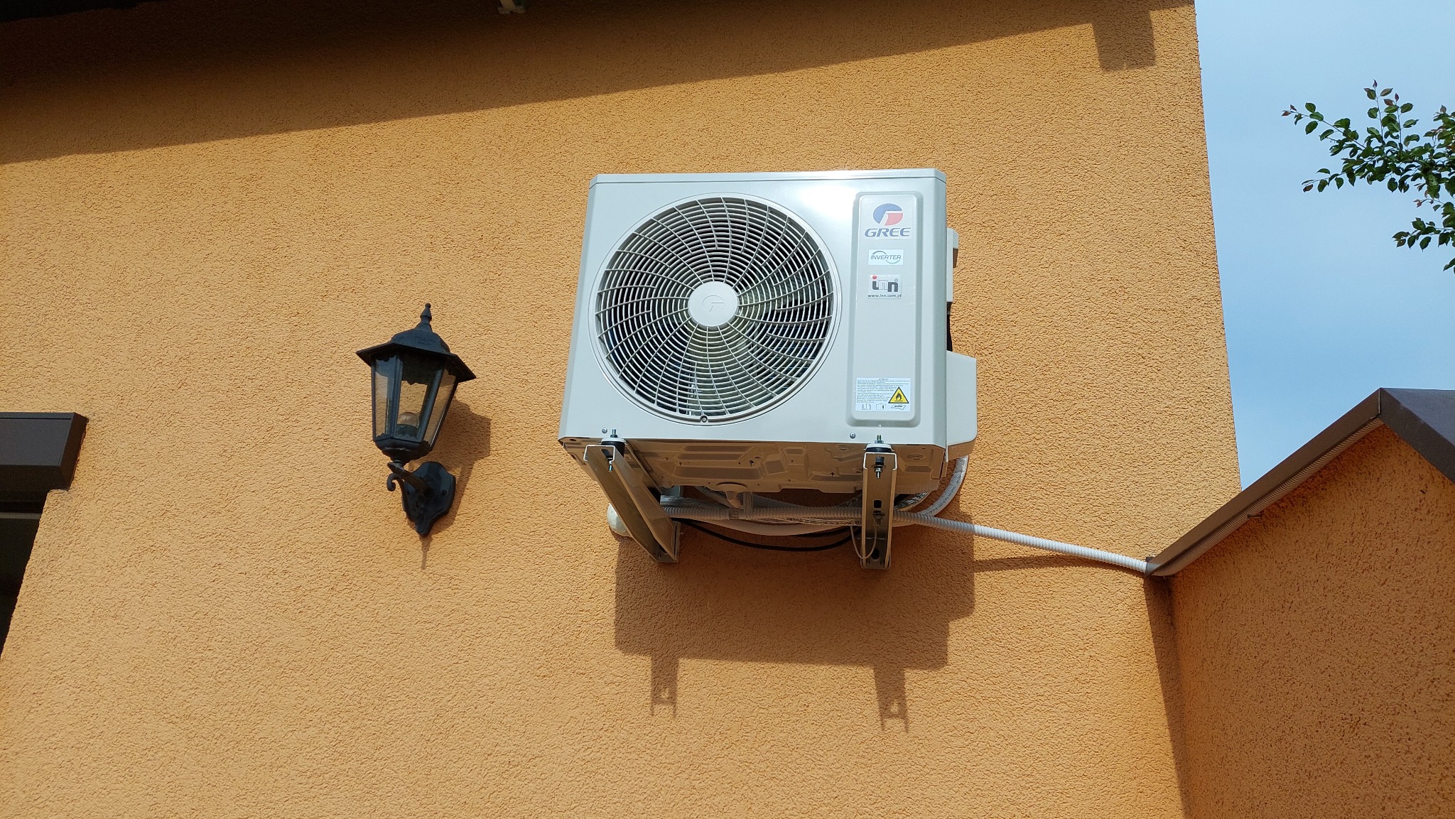 Zewnętrzna jednostka klimatyzacji marki Gree zamontowana na pomarańczowej, teksturowanej ścianie budynku, obok czarnej lampy ściennej.