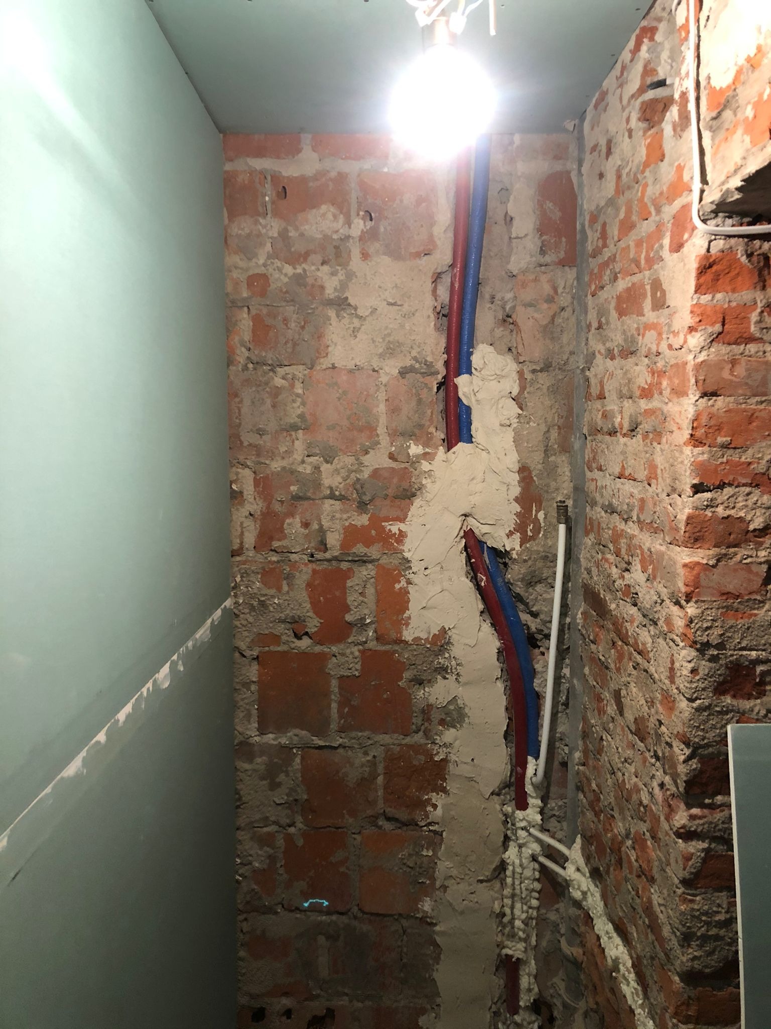 Surowa ściana z czerwonej cegły, częściowo pokryta gładzią, z wystającymi rurami wodnymi w kolorze czerwonym i niebieskim, oraz fragmentem płyty gipsowo-kartonowej na ścianie obok, oświetlona...