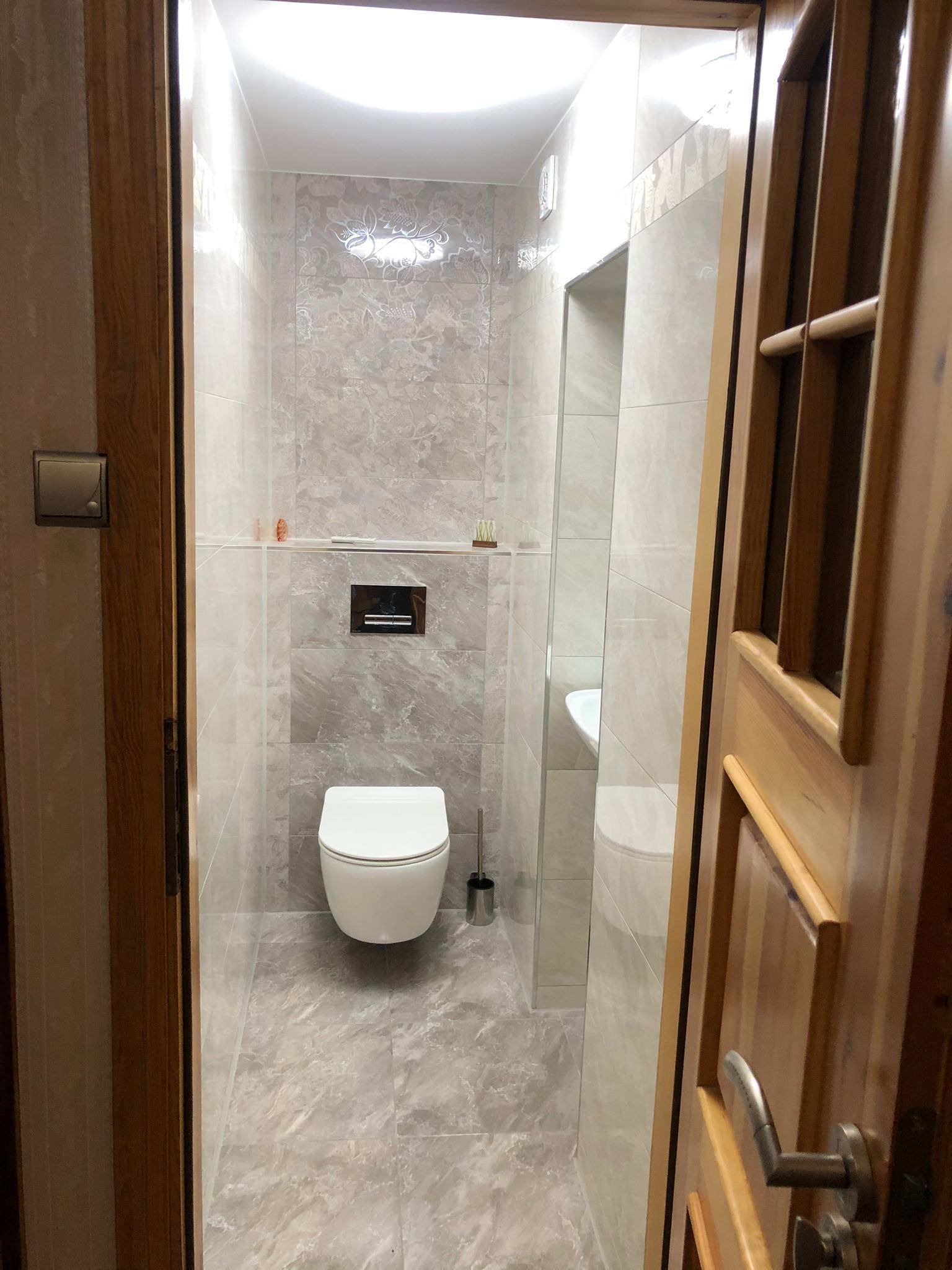 Nowoczesna łazienka z podwieszaną toaletą, wykończona płytkami imitującymi marmur, widoczna zza częściowo otwartych drewnianych drzwi z metalową klamką.