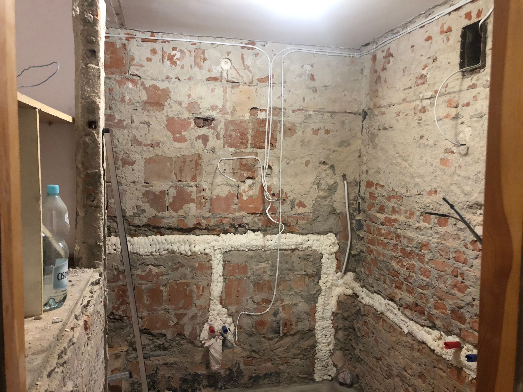Surowa ściana z cegły i resztkami tynku w trakcie prac instalacyjnych w łazience, z widocznymi przewodami elektrycznymi, rurami zabezpieczonymi pianką montażową, oraz butelką wody w tle.