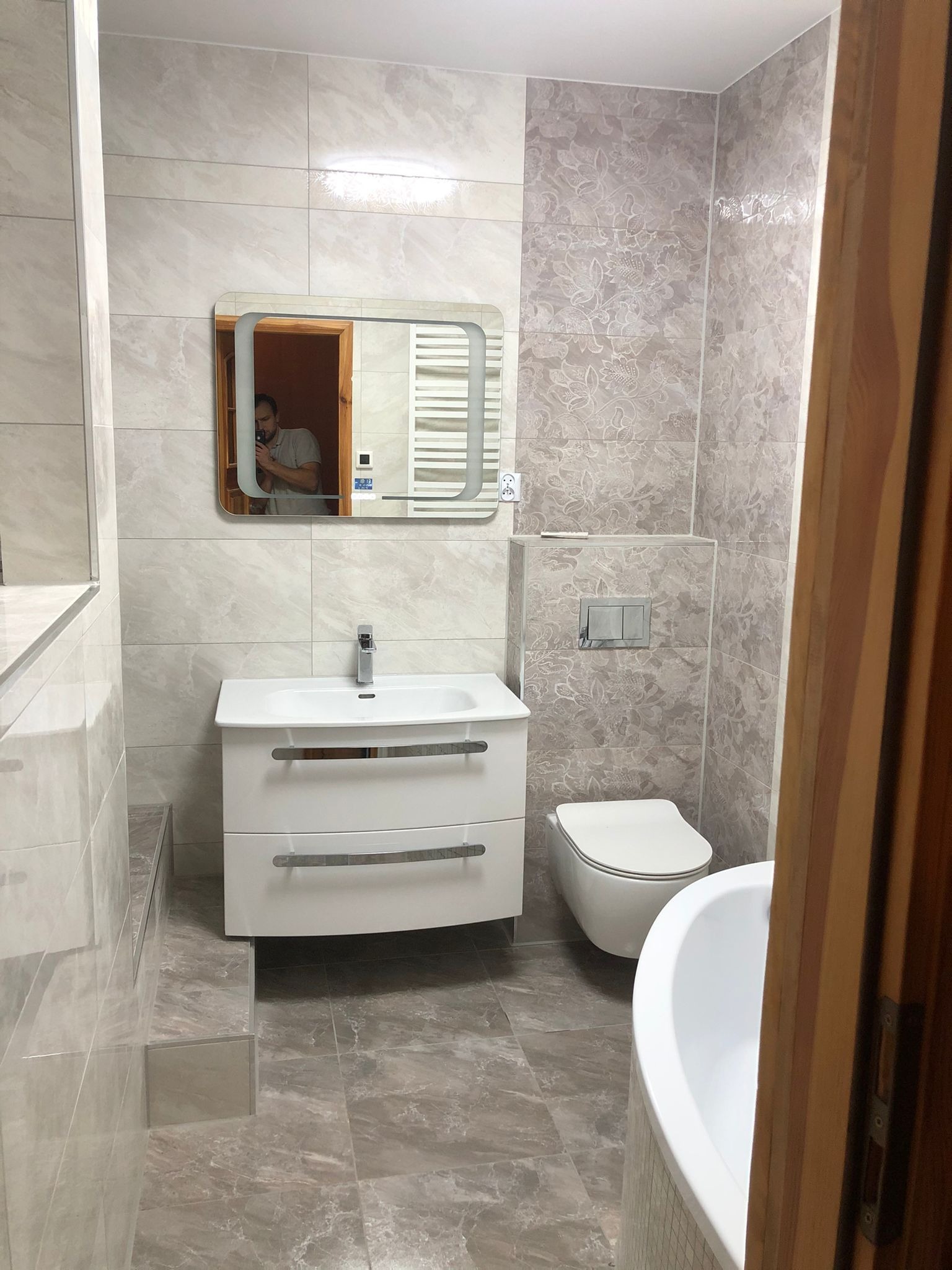 Nowoczesna łazienka z białymi sanitariatami, umywalką z szafką i podświetlanym lustrem, wykończona płytkami w odcieniach beżu i szarości, z widocznym odbiciem osoby robiącej zdjęcie.