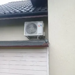 Biała jednostka zewnętrzna klimatyzacji Sinclair zamontowana na elewacji budynku, widoczna z boku, z elementami instalacji i czujnikiem na dachu garażu.