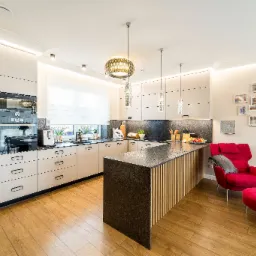 Nowoczesna kuchnia z wyspą, drewnianą podłogą i akcentem w postaci czerwonego fotela z podnóżkiem. Ściana ozdobiona galerią ramek ze zdjęciami.