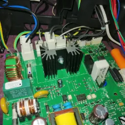 Zielona płytka drukowana z elementami elektronicznymi, radiatorami i przewodami w zbliżeniu, widoczne kondensatory, rezystory i układy scalone.