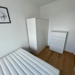 Minimalistyczna sypialnia z łóżkiem z materacem w białe pasy, białą szafą dwudrzwiową i komodą z czterema szufladami na drewnianej podłodze, ozdobiona abstrakcyjnym obrazem w czarnej ramie.