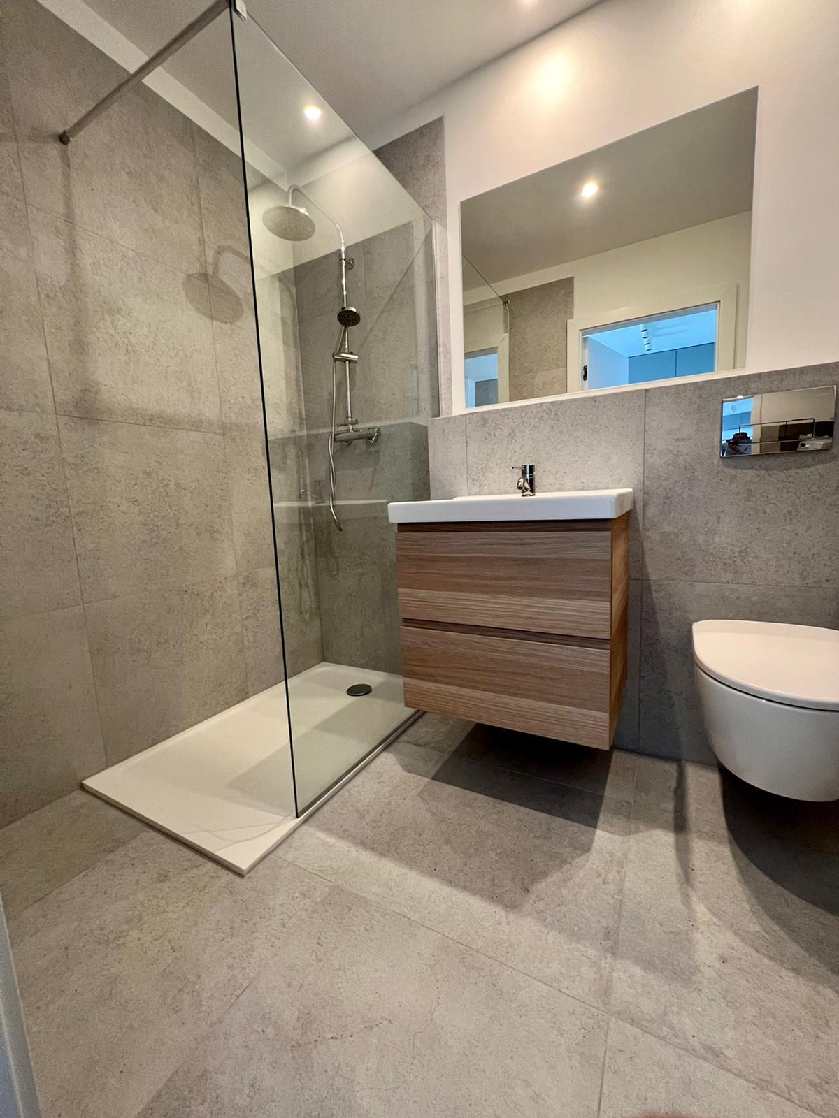 Nowoczesna łazienka z kabiną prysznicową typu walk-in, podwieszaną szafką z umywalką i toaletą w kolorze białym, wykończona szarymi płytkami.