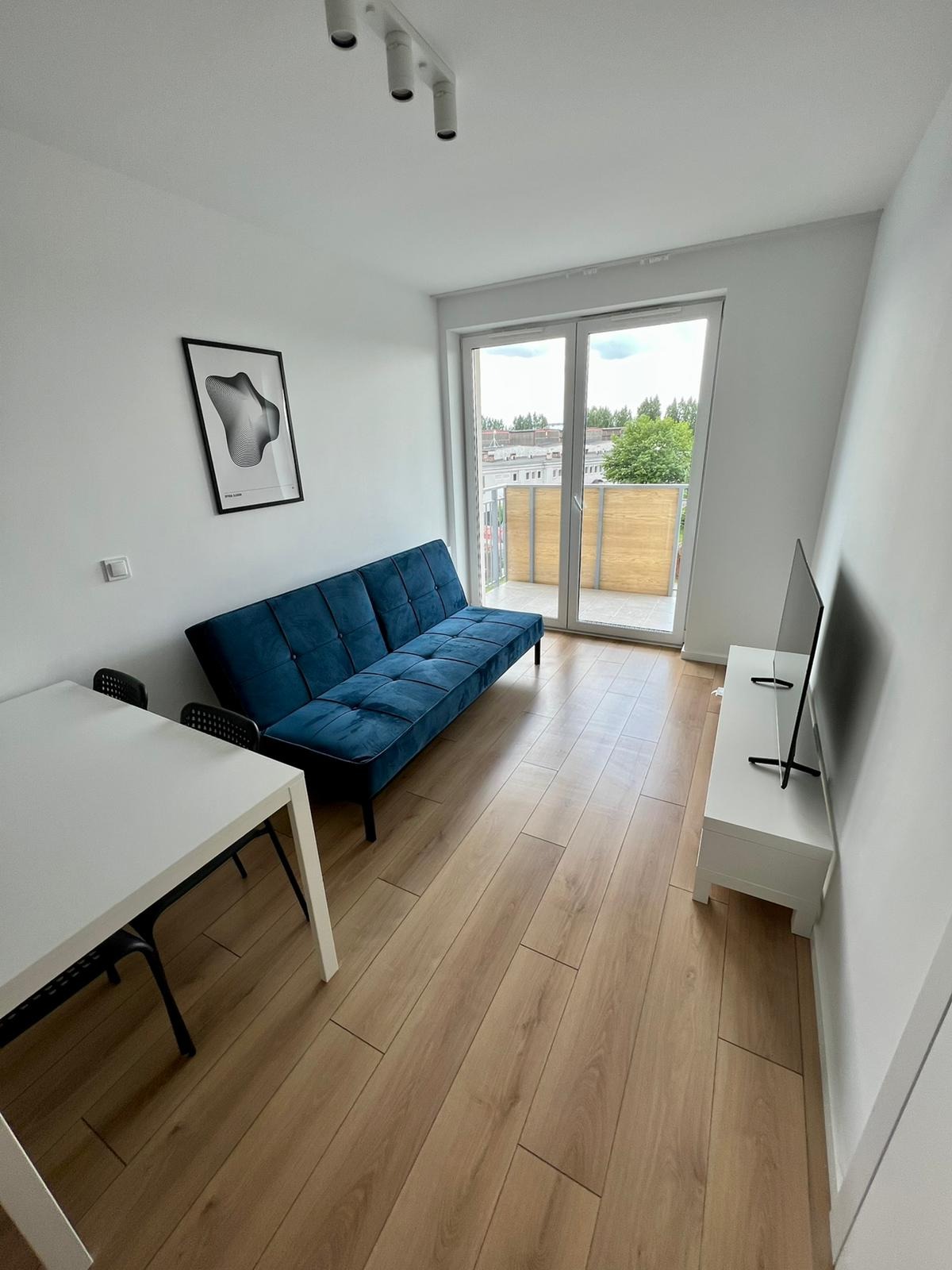 Jasne wnętrze pokoju z drewnianą podłogą, niebieską sofą, białym stołem z czarnymi krzesłami, szafką RTV z telewizorem i wyjściem na balkon.