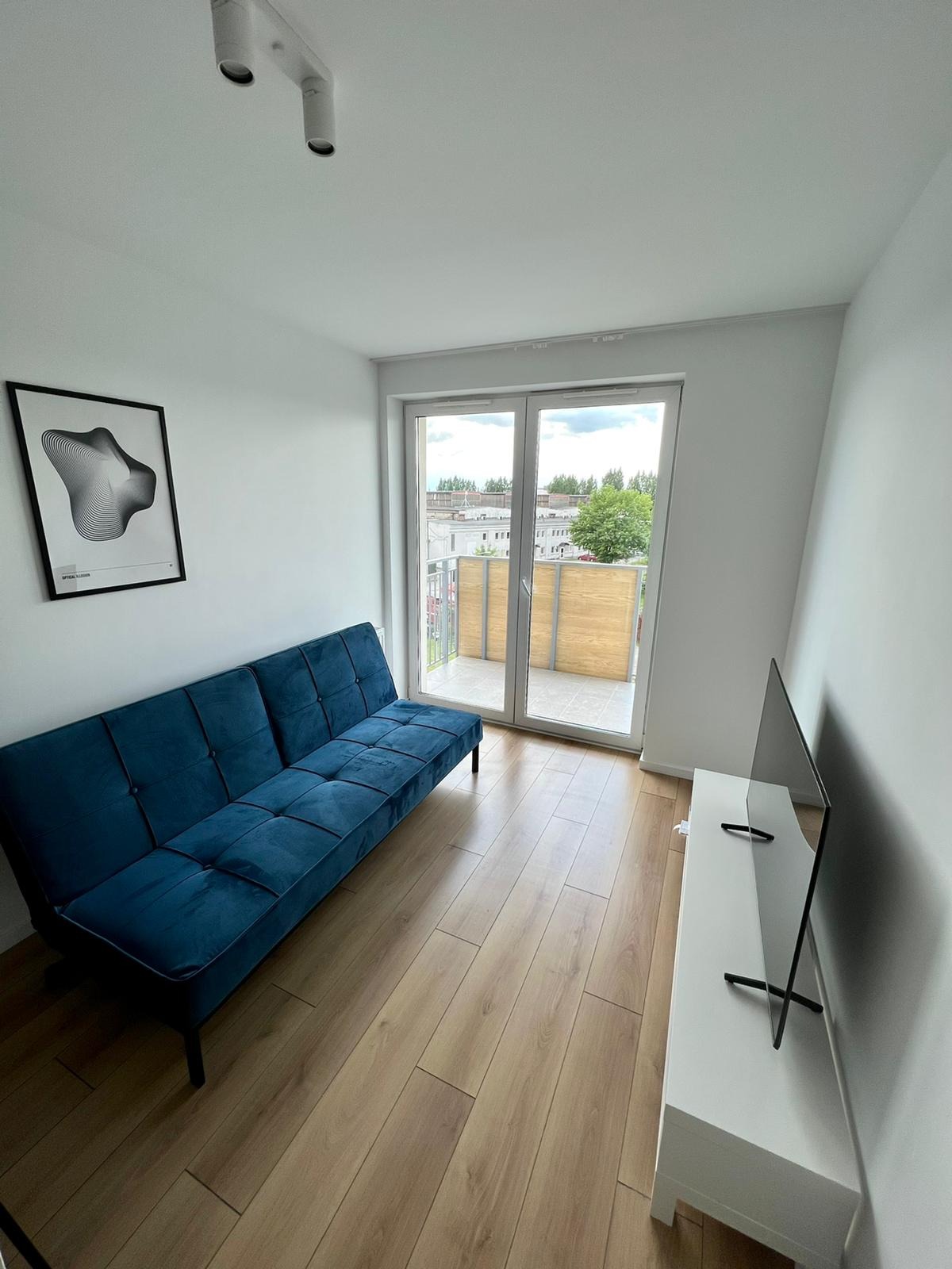 Jasne wnętrze pokoju z rozkładaną, turkusową sofą, telewizorem na białej szafce RTV i wyjściem na balkon z drewnianą balustradą, uzupełnione laminowaną podłogą w odcieniach drewna...