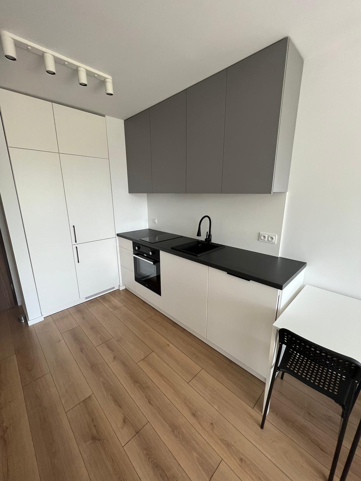 Nowoczesna kuchnia w odcieniach bieli i szarości z czarnymi akcentami, drewniana podłoga, minimalistyczny design, oświetlenie sufitowe punktowe.