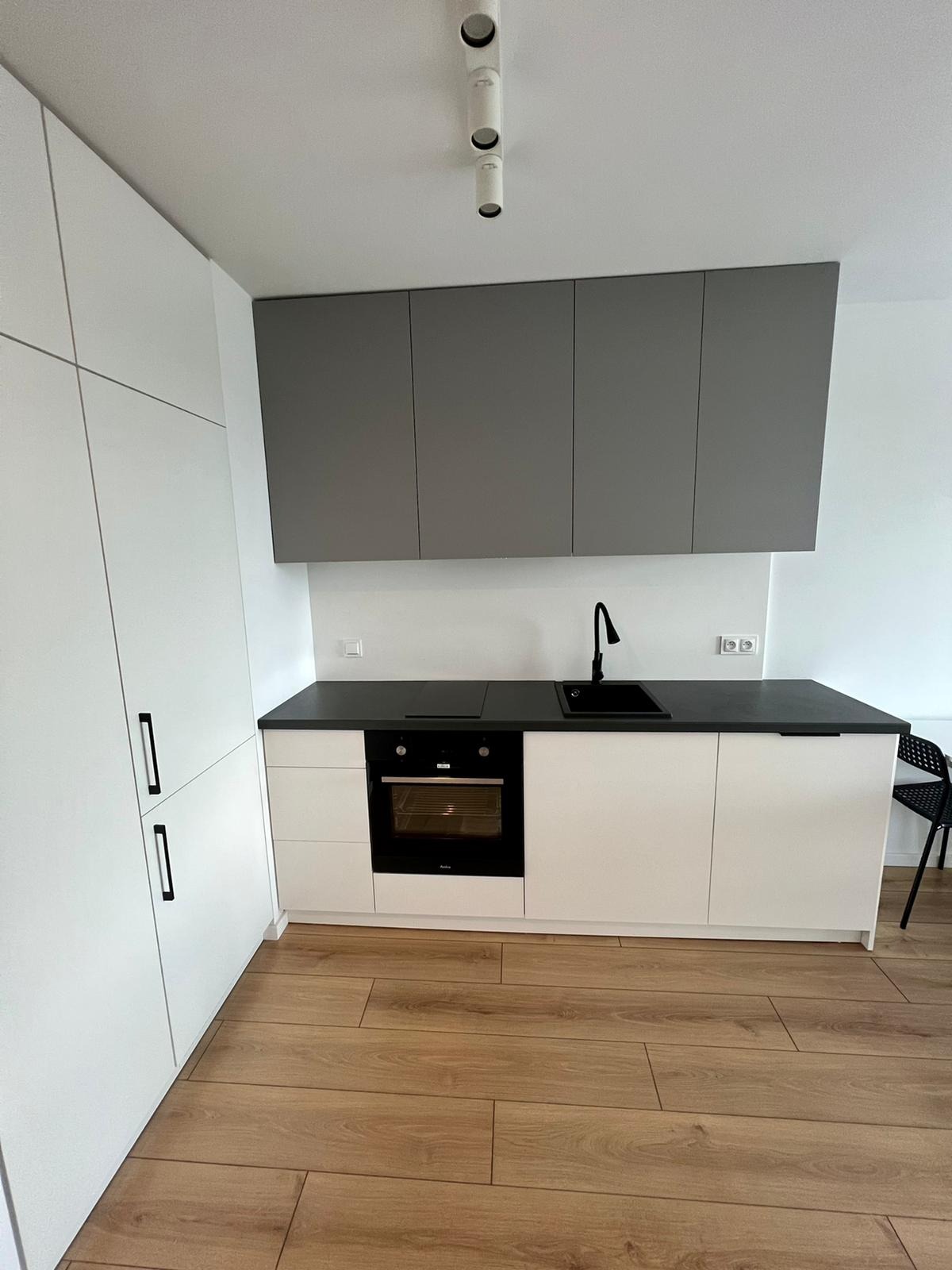 Nowoczesna kuchnia z białymi szafkami dolnymi i szarymi górnymi, czarnym blatem i armaturą, oraz drewnianą podłogą. Widoczny piekarnik Amica.