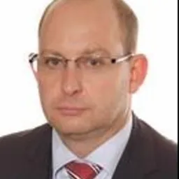 Portret mężczyzny w okularach, w garniturze i krawacie w odcieniach czerwieni, na jednolitym tle.