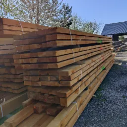 Stos tarcicy budowlanej z widocznymi oznaczeniami kolorystycznymi na końcach, zabezpieczony białymi taśmami, na tle żwirowego podłoża i budynków gospodarczych.