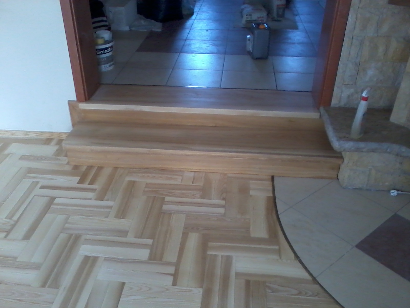 Parkiet jesionowy ułożony we wzór jodełki francuskiej, przechodzący w schodek z jasnego drewna i płytki podłogowe w przejściu do innego pomieszczenia.