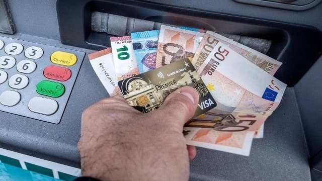 Ręka trzymająca kartę kredytową Visa Premier oraz banknoty euro przed bankomatem.