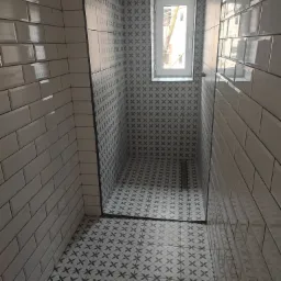 Nowoczesna kabina prysznicowa wyłożona białymi, błyszczącymi płytkami typu metro oraz wzorzystymi płytkami w odcieniach szarości, z odpływem liniowym i małym oknem w głębi.