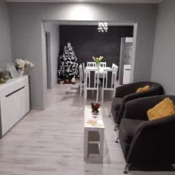 Nowoczesne wnętrze salonu z szarymi ścianami, jasną podłogą panelową, ciemnymi fotelami z żółtymi poduszkami i widokiem na jadalnię z choinką w tle.