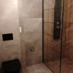 Nowoczesna łazienka z szarymi płytkami imitującymi marmur, czarną toaletą wiszącą i kabiną prysznicową typu walk-in z czarnymi profilami.