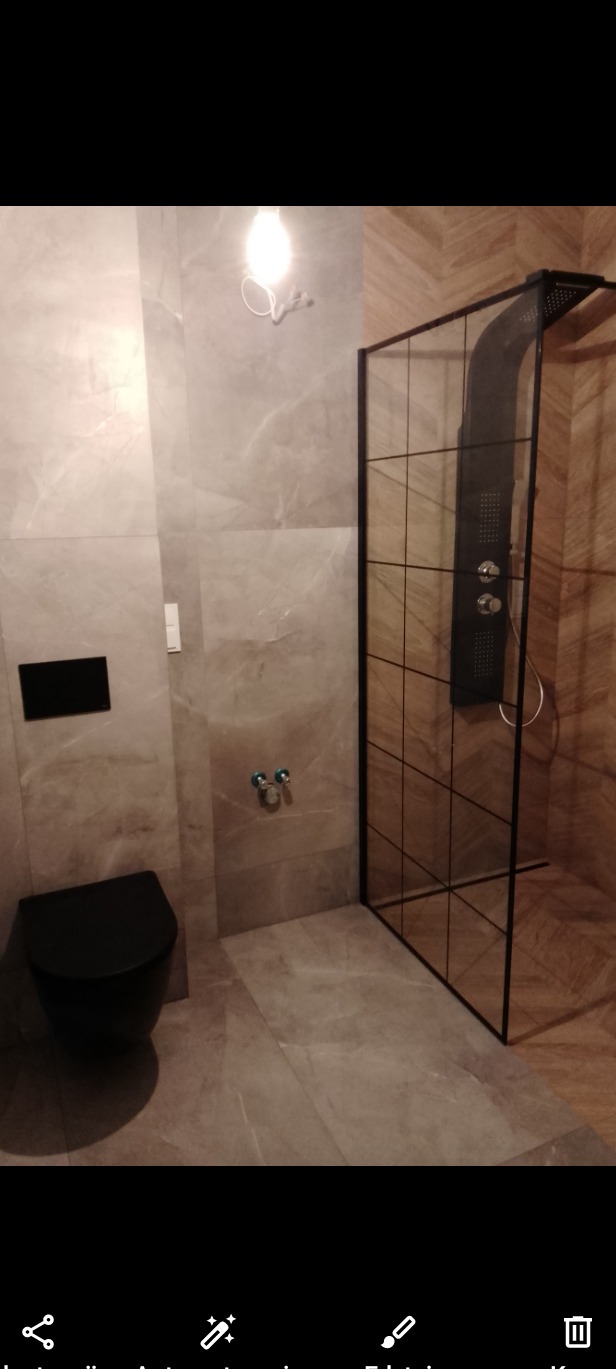 Nowoczesna łazienka z szarymi płytkami imitującymi marmur, czarną toaletą wiszącą i kabiną prysznicową typu walk-in z czarnymi profilami.