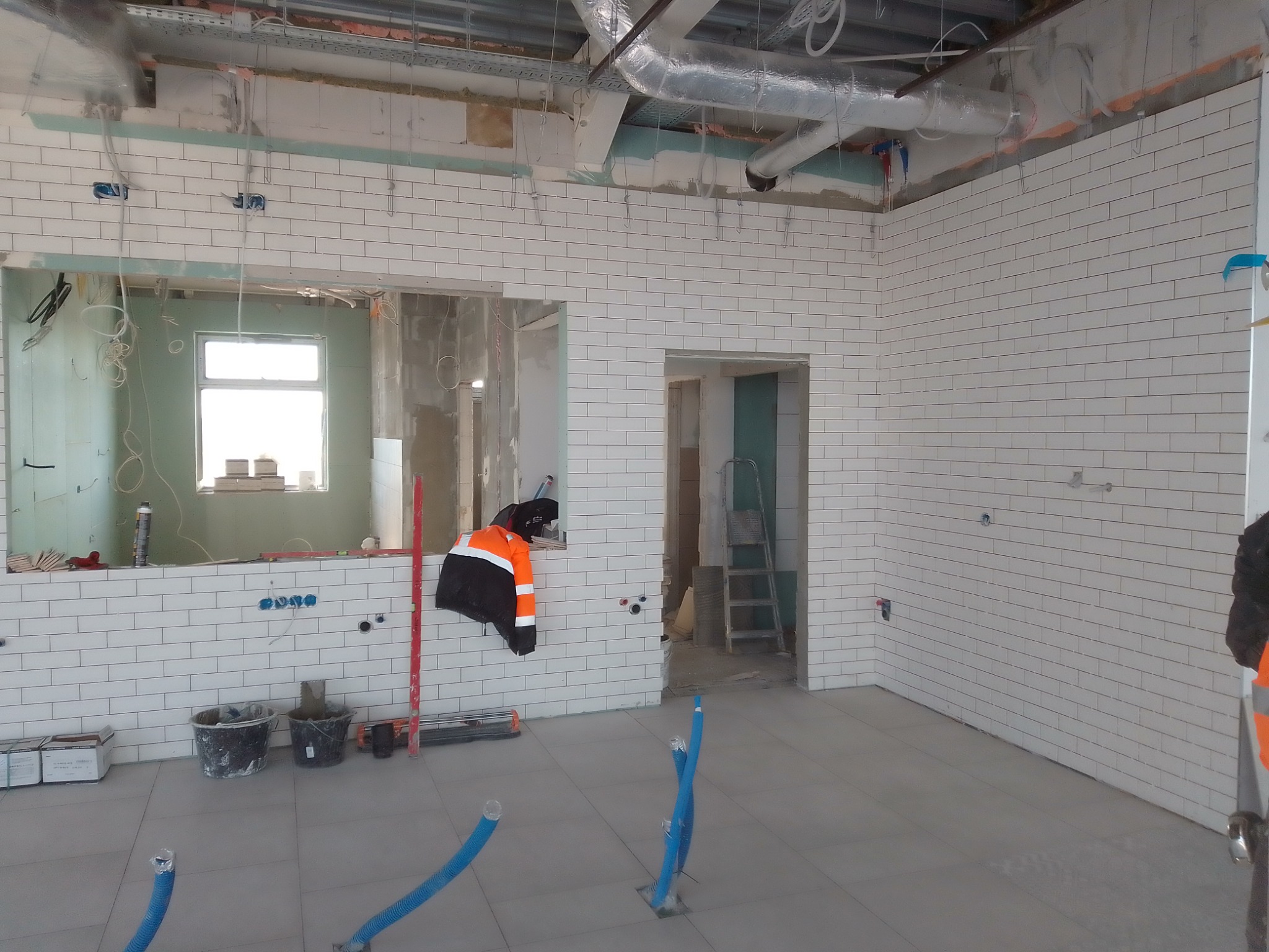 Wnętrze w trakcie remontu: białe płytki ułożone na ścianach, widoczne instalacje elektryczne, narzędzia, wiadro z zaprawą i odzież robocza