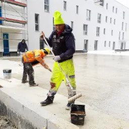 Dwóch pracowników w odblaskowych ubraniach maluje posadzkę przemysłową wałkami na zewnątrz budynku w trakcie budowy.