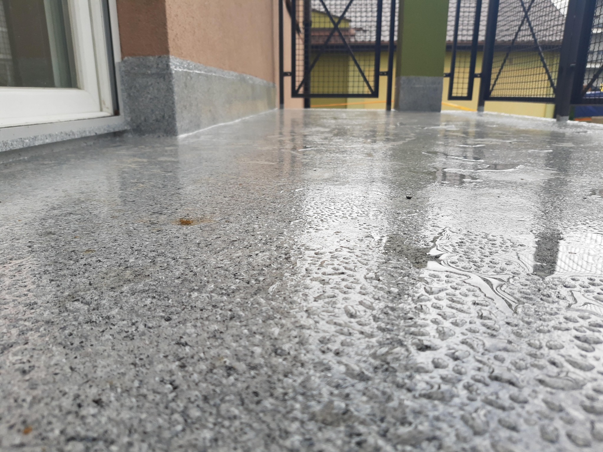 Szara, mokra posadzka na balkonie, z widocznymi kroplami wody i odbiciami metalowej balustrady. Widoczny fragment ściany budynku i okno.