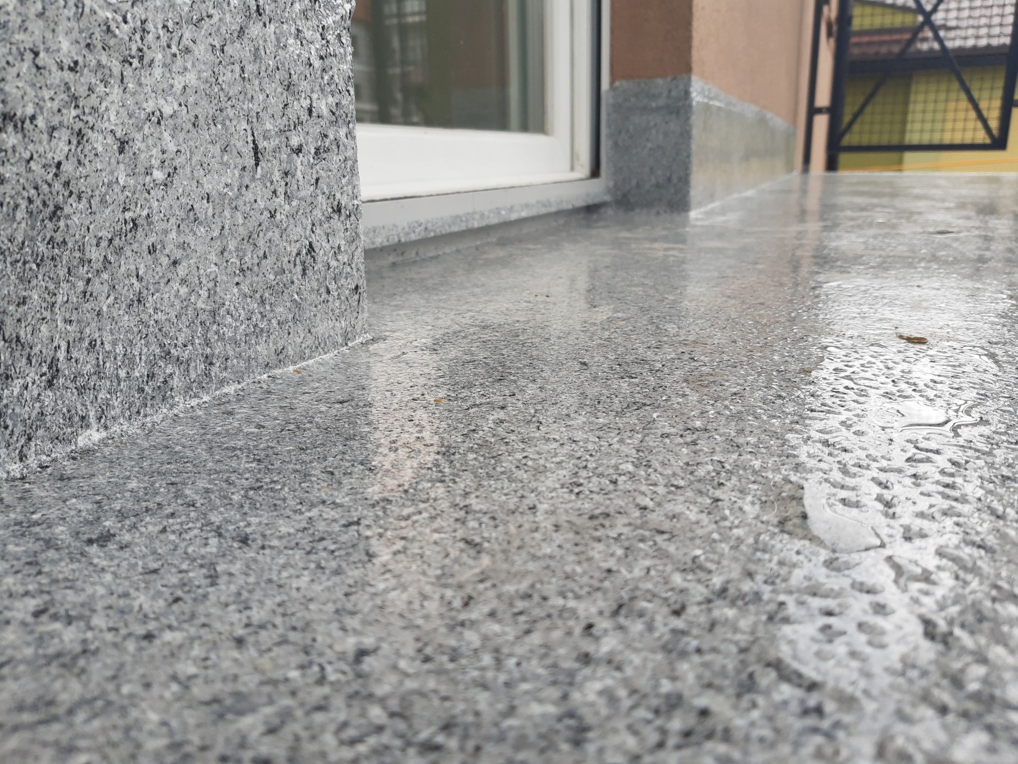 Szara, mokra nawierzchnia balkonu z widoczną teksturą kamienia, przylegająca do ściany budynku z szarą okładziną. W tle widoczna brama i fragment elewacji.